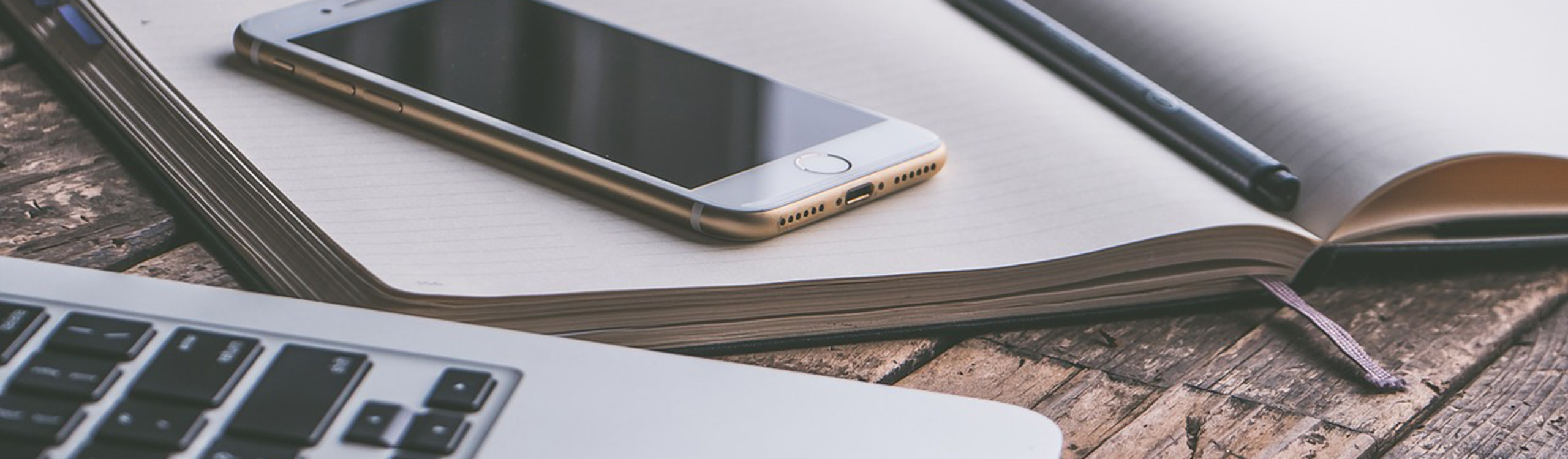 ein Smartphone liegt auf einem geöffneten Notizbuch neben einem Stift und einem Laptop, welche sich auf einem Holztisch befinden