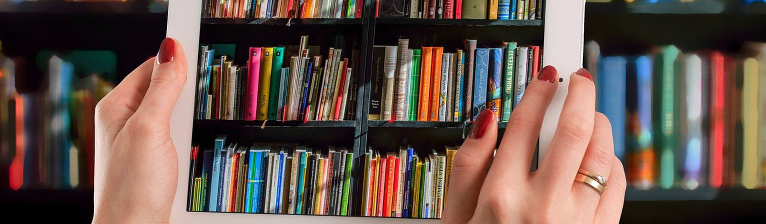 Hände halten ein Tablet, das ein Foto von einem Bücherregal macht, im Hintergrund sind die unscharfen Bücherregale zu sehen